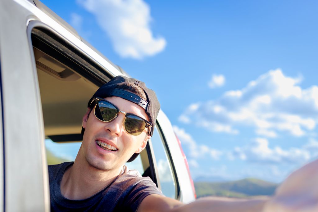 Un jeune homme souriant avec un appareil dentaire prend un selfie depuis une voiture, évoquant la confiance en son sourire.